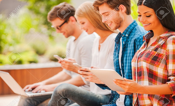 Digital world. Group of happy young people holding different digital devices and smiling while sitting in a row outdoors